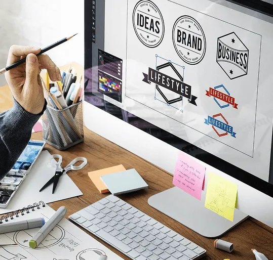 Espacio de diseño con conceptos de logos en pantalla y herramientas de boceto en el escritorio.